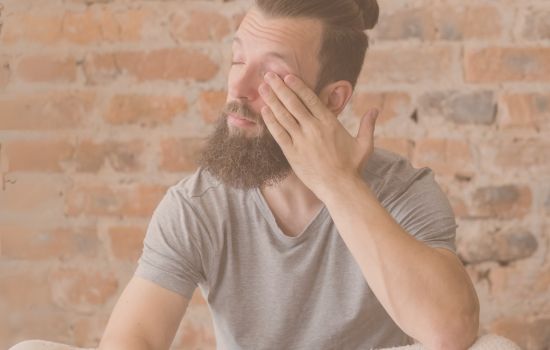 A men 30+ sitting and facing to his right side with closed eyes and he is rubbing his left eye with his left hand.
